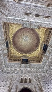 Alcázar de Sevilla features a domed, gold-adorned ceiling, intricate wall carvings, and small black-railed balconies.