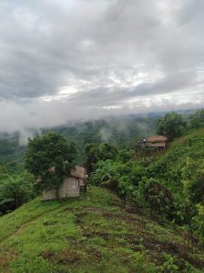 A small, simple hut with a tin roof sits on a green hillside in Lama, Bandarban, Bangladesh, with a larger, thatched-roof structure visible further up the hill. The surrounding hills are covered in thick green foliage, and a thick layer of fog hangs over the valley below, partially obscuring the distant mountains under a cloudy, overcast sky.