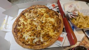 A large pizza topped with chunks of grilled chicken and melted cheese sits in a pizza box. There are several packets of sauce nearby, along with a portion of French fries in a separate container.
