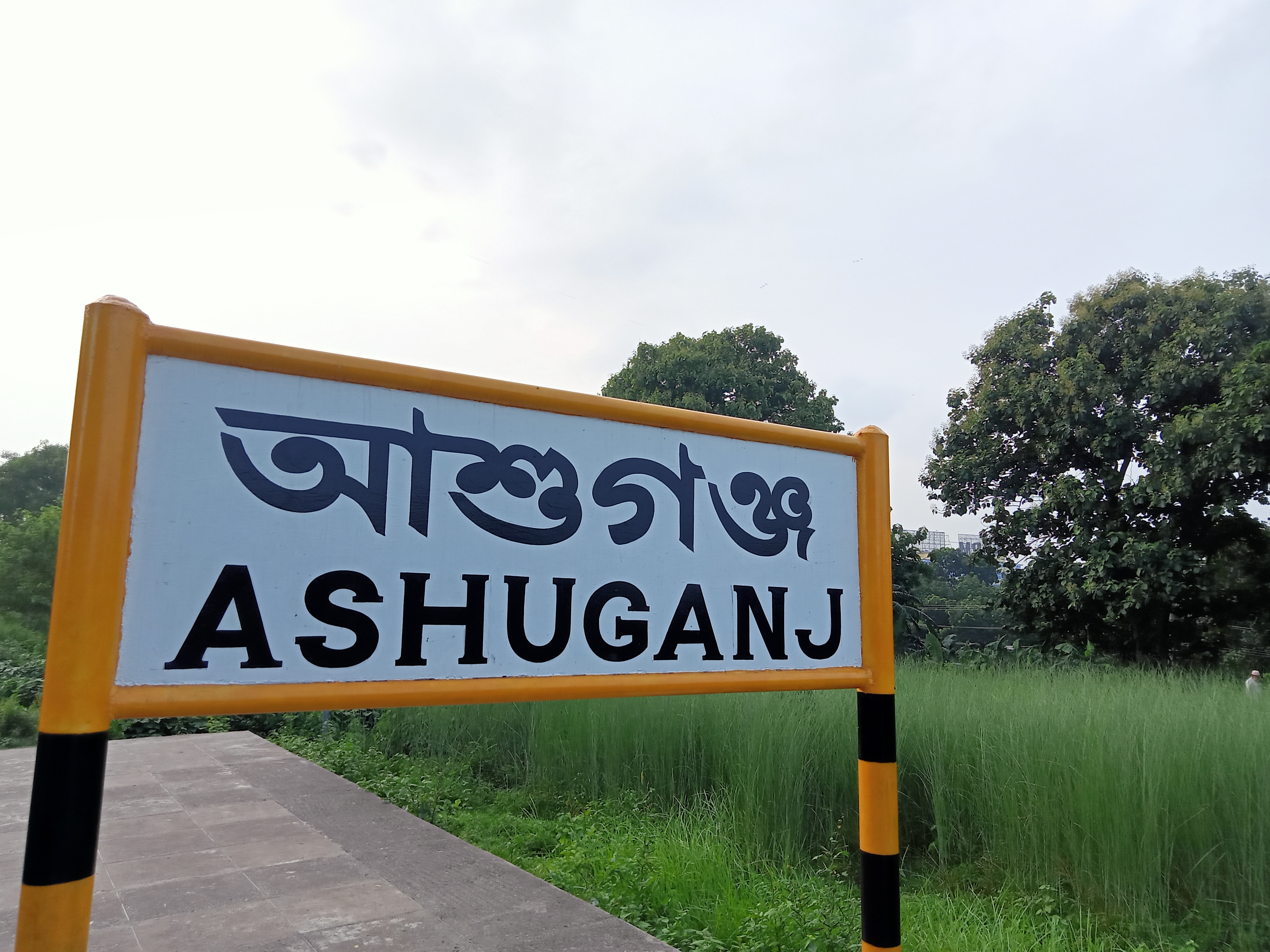 A signpost indicating the name "ASHUGANJ" in both Bengali script and English, with a background of lush green grass and trees under a cloudy sky.