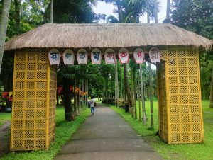 A thatched entrance gate made of bamboo with decorative panels displaying colorful text in a language that is not English. The gate stands in a green park surrounded by tall palm trees and a pathway leading into the greenery. A person can be seen walking along the path, which is lined with colorful flags.