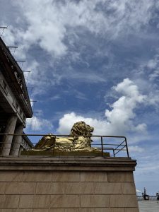 A golden lion statue on a stone platform with a railing, beside a classical columned building under a blue sky with white clouds.