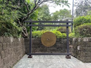 A brass gong on a wooden frame stands outdoors against a stone wall and greenery