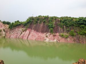 A natural landscape featuring a steep, rocky cliff with reddish hues, partially covered in greenery, adjacent to a river.