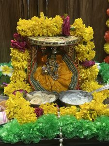 A beautifully decorated idol of Lord Krishna, dressed in vibrant yellow attire, adorned with marigold garlands, and surrounded by offerings of food neatly placed in bowls.
