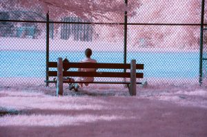 A person sits alone on a park bench facing a chain-link fence and baseball diamond. Captured in surreal colors from infrared photography. 
