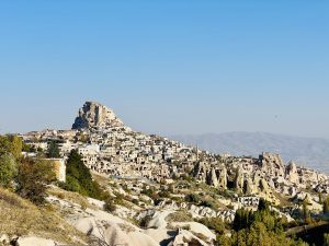 A wide view of a village with homes carved into a hillside of rock formations under a clear blue sky, at Cappadocia, Turkey.