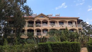 A nice guest house in Uganda. A three-story building with a pink and beige facade is surrounded by lush greenery and plants. The building features several balconies with white railings and plants growing along the edges.