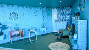 The blue room at the TWA hotel at JFK Airport. A vignette of a blue midcentury living room with blue shag carpet, blue walls, blue padded mini bar, a blue brick fireplace, and blue curtains with geometric patterns.