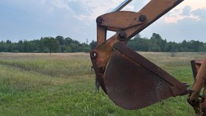 in the foreground is the bucket of a backhoe, a large open field in the background.  The bucket is old and rusty.