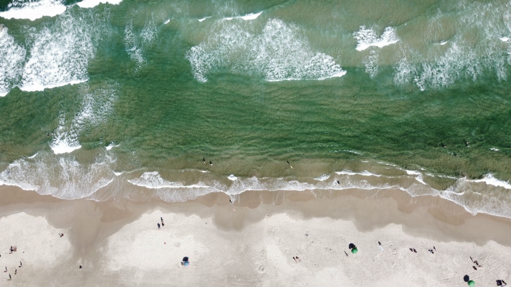 An aerial view of a beach with gentle waves lapping at the shore. People can be seen swimming and surfing in the water. On the sandy beach, there are small groups of beachgoers relaxing, with some umbrellas set up.