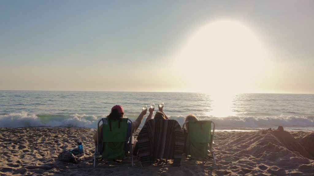 3 best friends sitting in chairs on a beach watching the sunset and raising a glass of wine to toast one another.