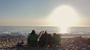 3 best friends sitting in chairs on a beach watching the sunset and raising a glass of wine to toast one another.