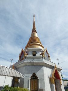 A low-angle shot of a large, gleaming golden stupa or pagoda in Thailand, set against a bright blue sky with scattered clouds.