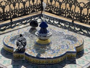 A sunlit fountain, top view. It's a low fountain, decorated with hand-painted tiles and bright colors. Water falls from the spout, and the sun shines in the drops below. Two pigeons are drinking from it, perched on the edge. In the background, the fence surrounding the fountain appears blurred.