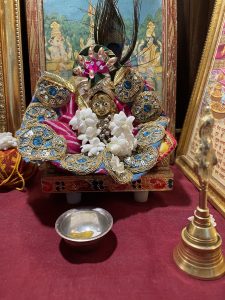 Lord Krishna adorned with colorful clothes, jewelry, and fresh white flowers, placed on a wooden pedestal with a red cloth background. A silver bowl and a brass bell are kept nearby, with devotional paintings in golden frames behind kanha.
