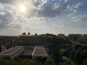 A picturesque landscape featuring rolling hills under a partly cloudy sky. The sun is peeking through the clouds, casting rays of light down. In the foreground, several low-rise buildings with flat roofs are visible alongside lush greenery. There are trees scattered on the hills in the background, with some terraced landscaping.
#SummerPhotoContes
