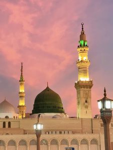 The Prophet's Mosque in Medina, Saudi Arabia at sunset, featuring a large green dome and tall minarets illuminated in golden light