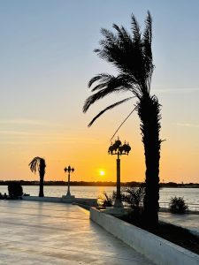 A picturesque sunset over the Jeddah Waterfront promenade. Silhouetted palm trees frame the scene, while elegant streetlamps stand against the vibrant orange and yellow sky.