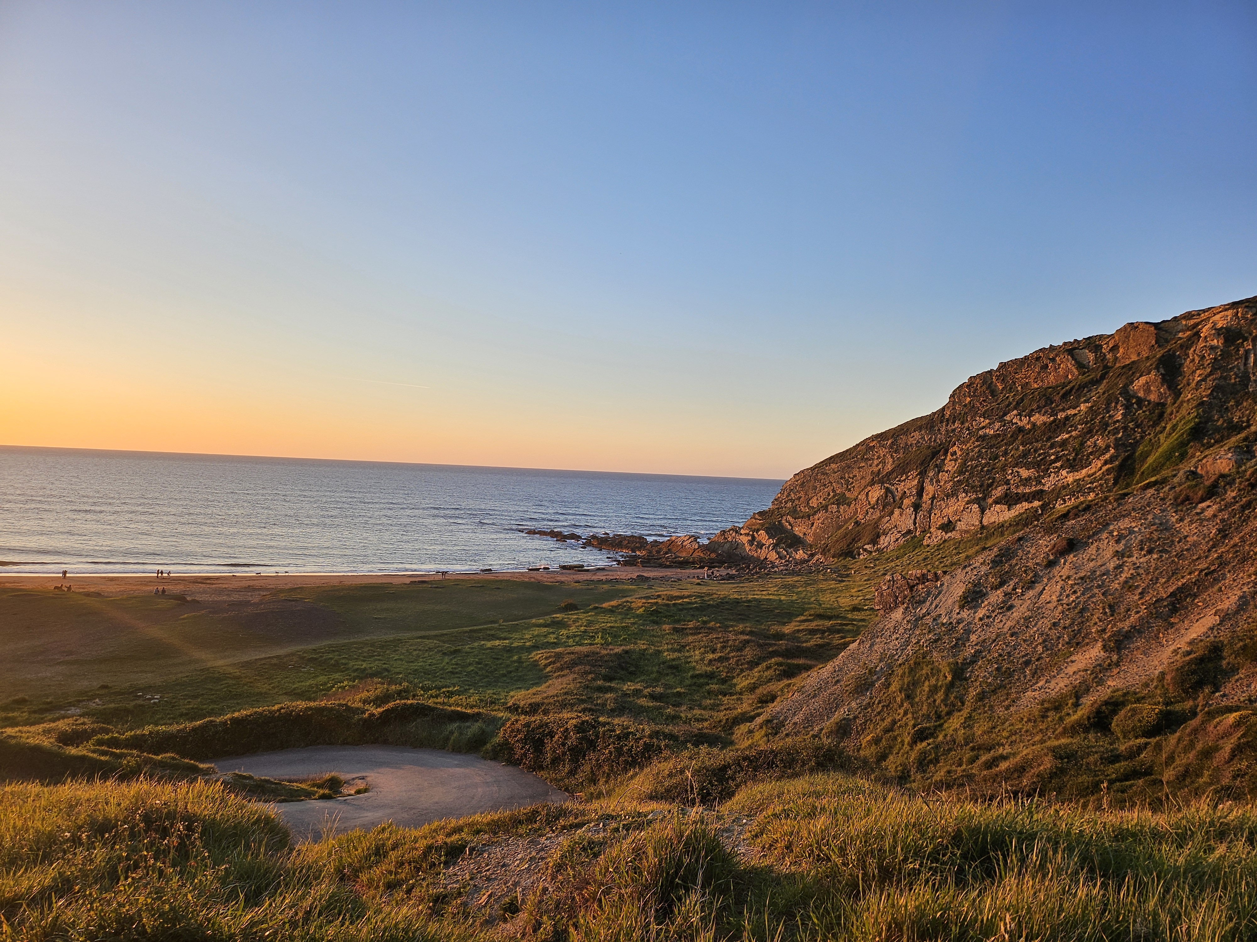 Panoramic view of a cliff covered in vegetation at sunset, with the sun casting a golden glow over the slopes and the sky in soft shades of blue and orange. Below, a quiet beach lies by the sea, with small figures of people walking in the distance. A natural scene that conveys calm, spaciousness, and beauty