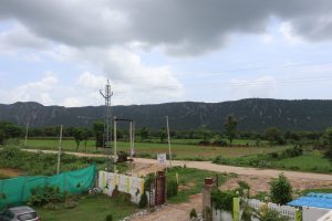 A view of Aravali Hills showing green hills and some people resting on the farming field with some trees. A man is going through the muddy road on his motorbike. Across the road seems a view of a house with cemented walls and a gate, with a car parked on its garden.