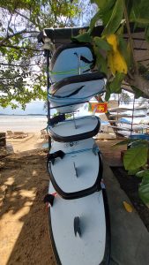 A rack of surfboards sits on a sandy beach under a tree, with the ocean visible in the background. Captured from Weligama Beach, Sri Lanka.