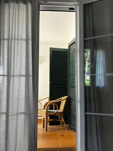 View through a terrace door: the left side closed with a curtain, the right side open onto two wicker chairs on a terrazzo floor, with a white wall and green shutters in the background.
