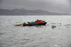 A rescue jet ski with orange and teal colors is floating on a calm sea, near a buoy. The background features a mountainous landscape under a cloudy sky, and the water reflects the overcast light.