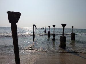 A wide-angle shot of a beach with old, rusted concrete pillars standing in the shallow water, with the sun reflecting off the ocean surface in the background.