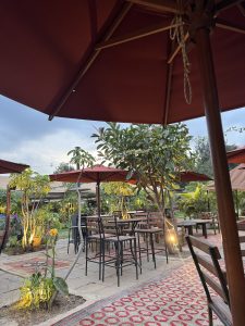 An outdoor summer patio with tables, chairs, red umbrellas under lush greenery