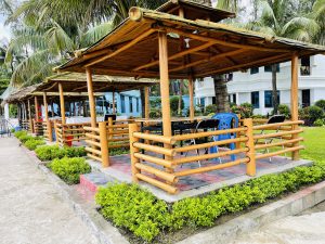 A series of small, open-air huts with bamboo roofs and railings, each with a table and chairs, set up in a lush green garden at a resort in Cox's Bazar.