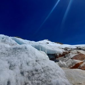 A close-up view of a snow and ice-covered mountain slope, with icy formations and a clear blue sky in the background. The image captures the textures of the snow and rocky surfaces, highlighting the contrast between the white ice and the reddish-brown rock. Sunlight reflects off the icy surfaces, creating a shimmering effect.