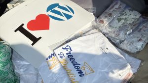 WordPress swag items displayed, including a large sign that says “I ♥ WordPress” with a blue WordPress logo next to it. There are white t-shirts folded and partially visible, one with the text “Freedom To Publish” accompanied by yellow and blue graphics. Surrounding the t-shirts, there are several packets containing stickers with various designs. The background features patterned fabric and some green packing material.
