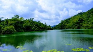 A tranquil view of Boga Lake in Bandarban, Bangladesh, surrounded by lush green hills under a partly cloudy sky.