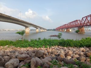 A picture of a two bridge going straight over a river and a wooden boat with few people passing underneath the bridges. Some oval and irregular shaped stones and green bushes are visible. Bhairab, Kishoreganj, Bangladesh #SummerPhotoContest