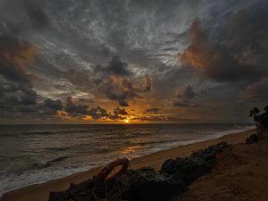A dramatic sunset over the ocean with vibrant orange and gold hues illuminating the clouds, set against a moody, darkened sky. The shoreline is visible, with gentle waves lapping at the sandy beach, and a lifebuoy resting on rocks in the foreground