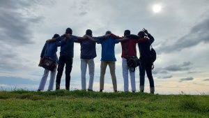 A low-angle shot from behind shows six young men standing in a line on a grassy hill, with their arms around each other's shoulders, facing a bright, cloudy sky. Captured from Gallery Fort, Sri Lanka. 