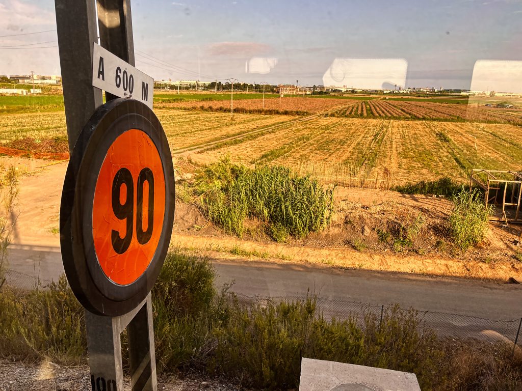 Through a train window with faint reflections, fields and distant buildings are visible under a slightly cloudy sky. On the left, a round orange train sign on a metal pole shows ’90’ for speed at 600 meters, with a narrow road running between the sign and the fields