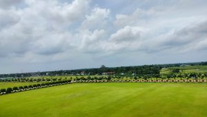 A panoramic view of a lush, green field bordered by neatly trimmed hedges and palm trees. In the background, a variety of structures, including low-rise buildings and a partially constructed structure, can be seen amidst a landscape of greenery under a cloudy sky. 
