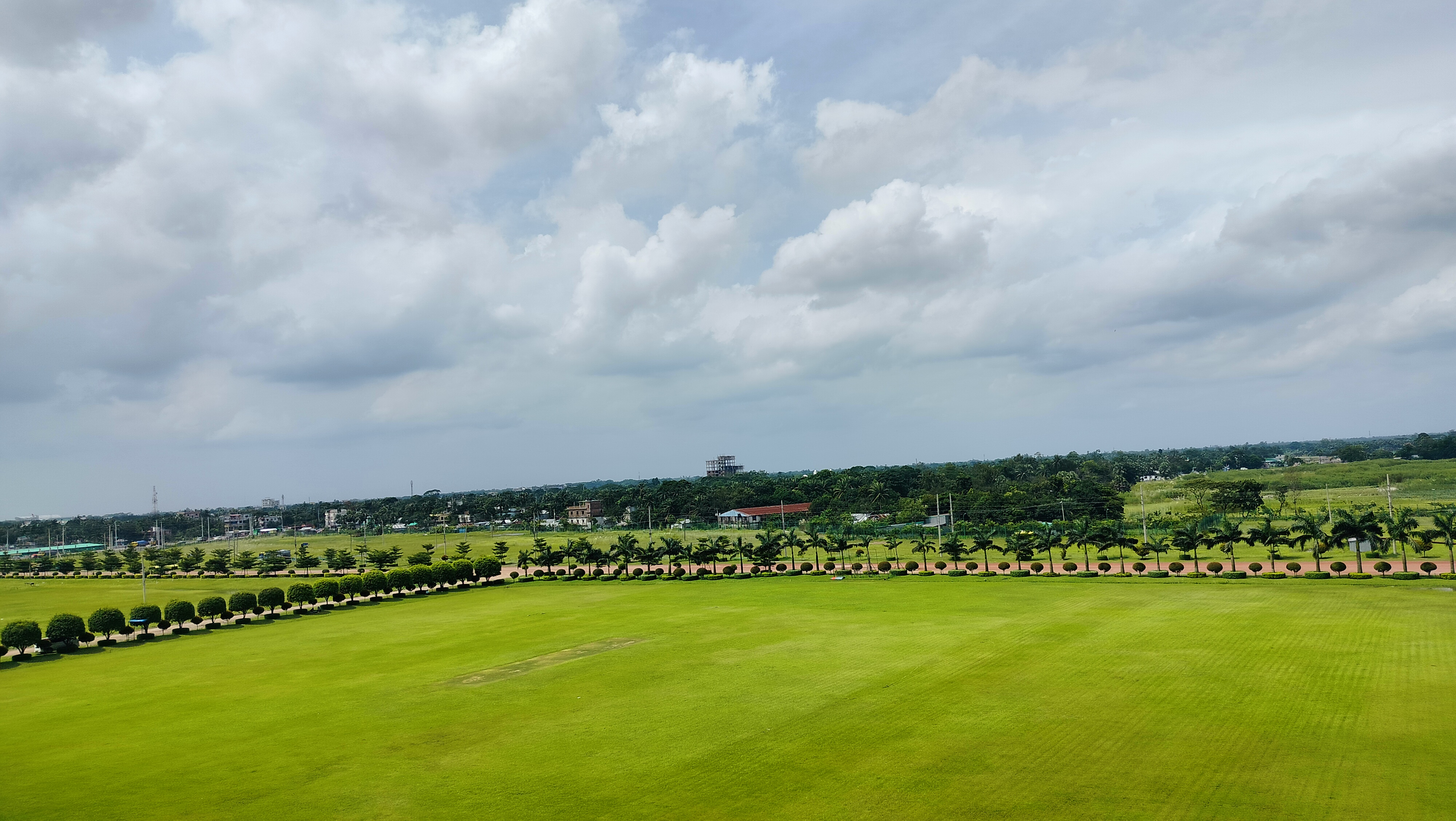 A panoramic view of a lush, green field bordered by neatly trimmed hedges and palm trees. In the background, a variety of structures, including low-rise buildings and a partially constructed structure, can be seen amidst a landscape of greenery under a cloudy sky.