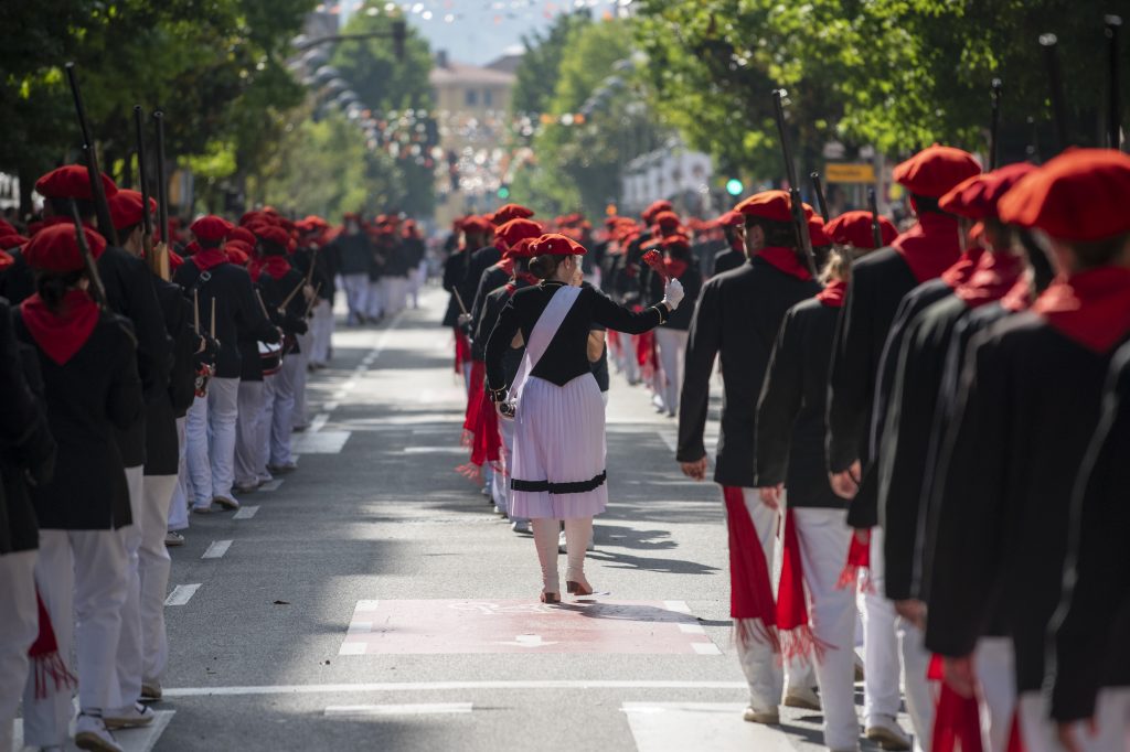 A woman dressed in traditional Basque attire, carrying a fan, leads two rows of participants wearing red berets, black jackets, and white trousers with red sashes during the Alarde de San Marcial parade in Irun, Spain