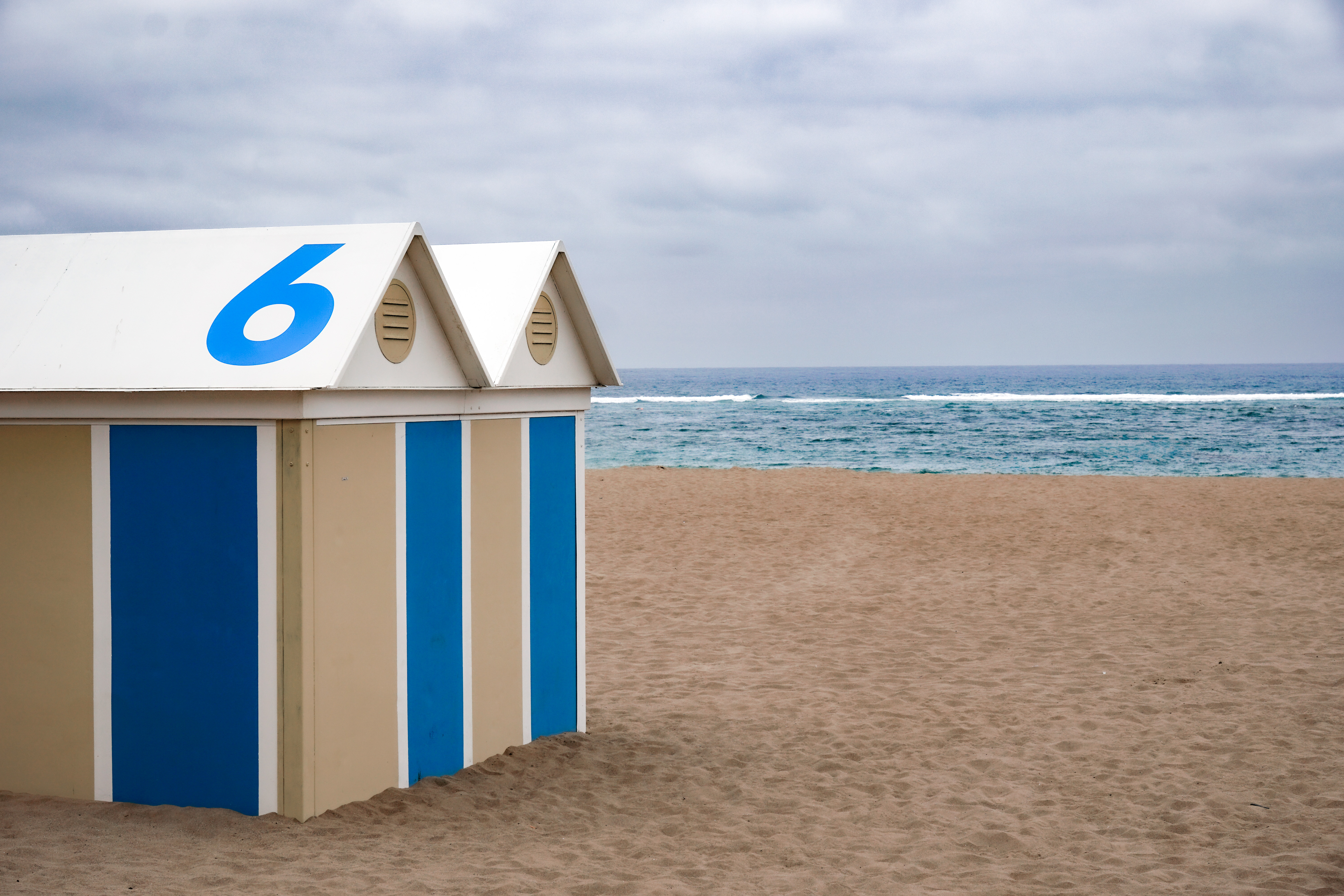 A beach scene featuring a beach hut with the number "6" on the roof. The hut has a light tan color with blue vertical stripes. In the background, there is a sandy beach leading down to the ocean, which has gentle waves under a cloudy sky.
