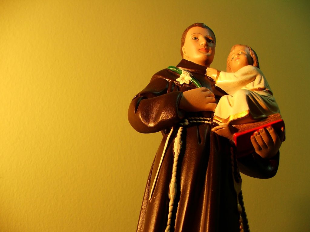 A close-up of a religious statue depicting Saint Anthony of Padua holding the infant Jesus. Saint Anthony is dressed in a brown Franciscan robe with a white cord belt, and the child is clothed in a white garment with golden accents, sitting on a red book. The background is a softly lit yellow wall, creating a warm and serene atmosphere.