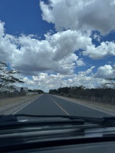 A first-person view from a car driving on a long, straight road. The road has a yellow dashed line down the middle and is bordered by dry, bushy land on either side. A bright blue sky filled with large, fluffy white clouds stretches overhead