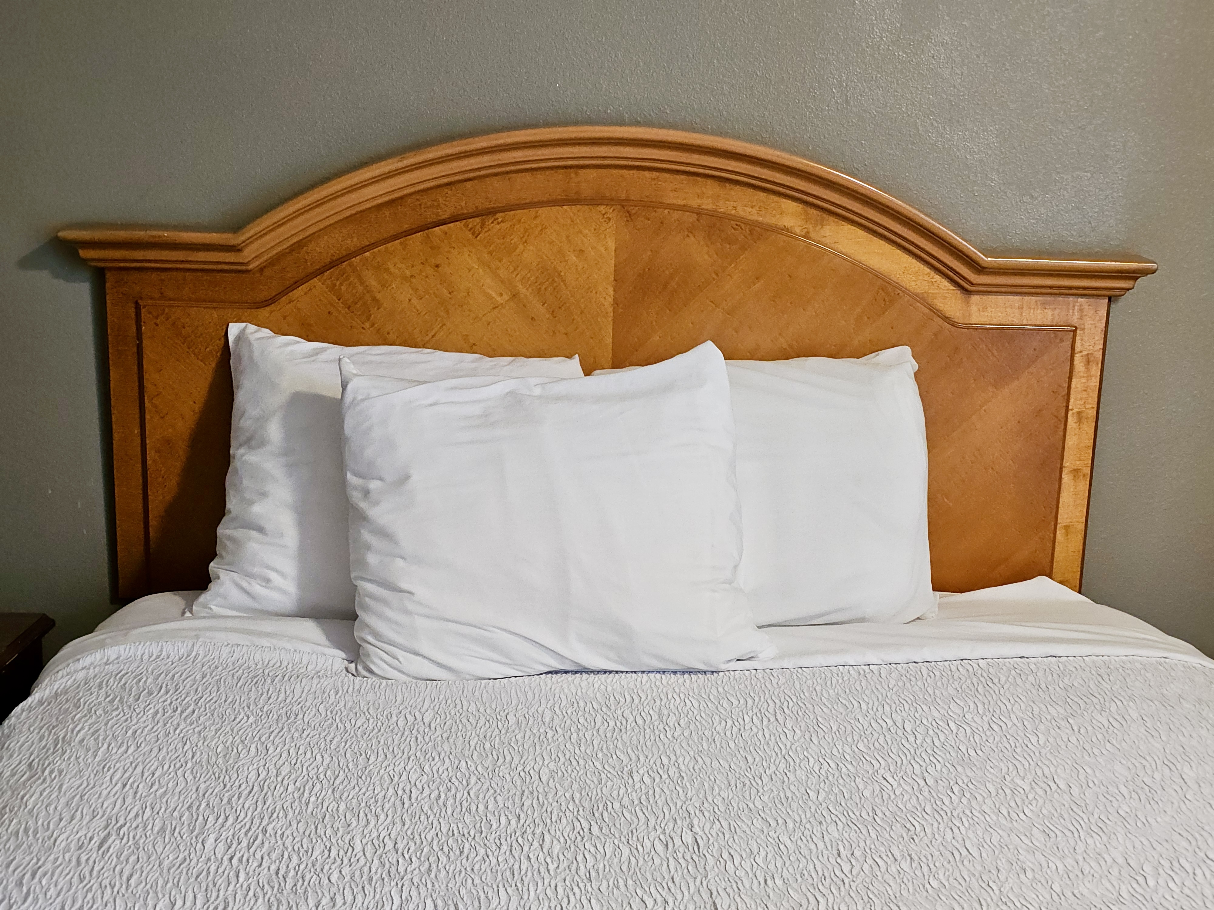 A hotel room, featuring a bed with a wooden headboard and white bedding, including a comforter and pillows.