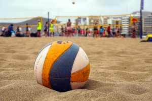 A close-up view of a volleyball resting on sandy beach terrain, with the blurred background showing a group of people engaged in a volleyball game at a beach court.