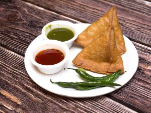  Two crispy golden samosas served on a white plate with green chilies and two small bowls of green chutney and red tamarind sauce, placed on a wooden table.
