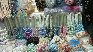 The image features a variety of decorative necklaces and jewelry displayed at a market stall. There are multiple strands of beads in different colors, shapes, and materials, including glass, pearls, and shells.