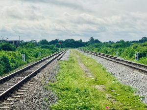 A view of two parallel railway tracks diverging in the distance, surrounded by lush green vegetation and scattered gravel. A small building can be seen on the left, and a red signal light is positioned near the tracks. The sky is partly cloudy, contributing to the overall natural landscape.