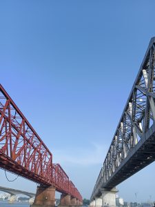 Bhairab, Kishoreganj, Bangladesh. Two lattice steel bridges—one red, one gray—run parallel over a river, converging in perspective beneath a clear blue sky. In the distance, another bridge, cranes, and buildings complete an industrial urban scene.